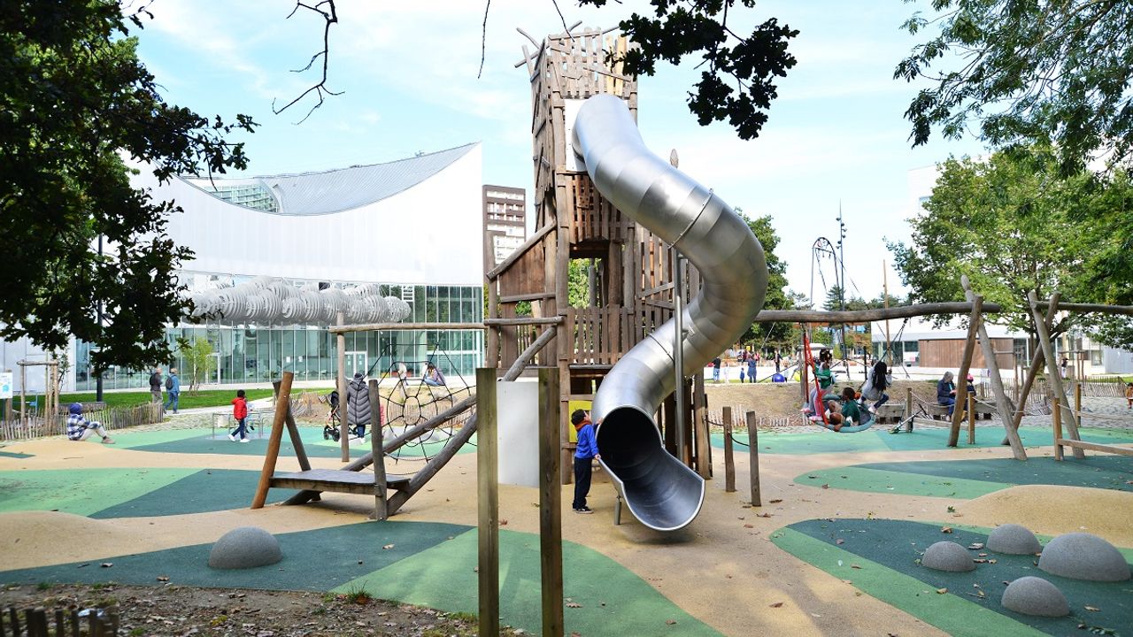 Vue des jeux pour enfants des Jardins Slovènes avec une structure avec un toboggan au premier plan, des enfants sur des balançoires et en arrière plan le Conservatoire.