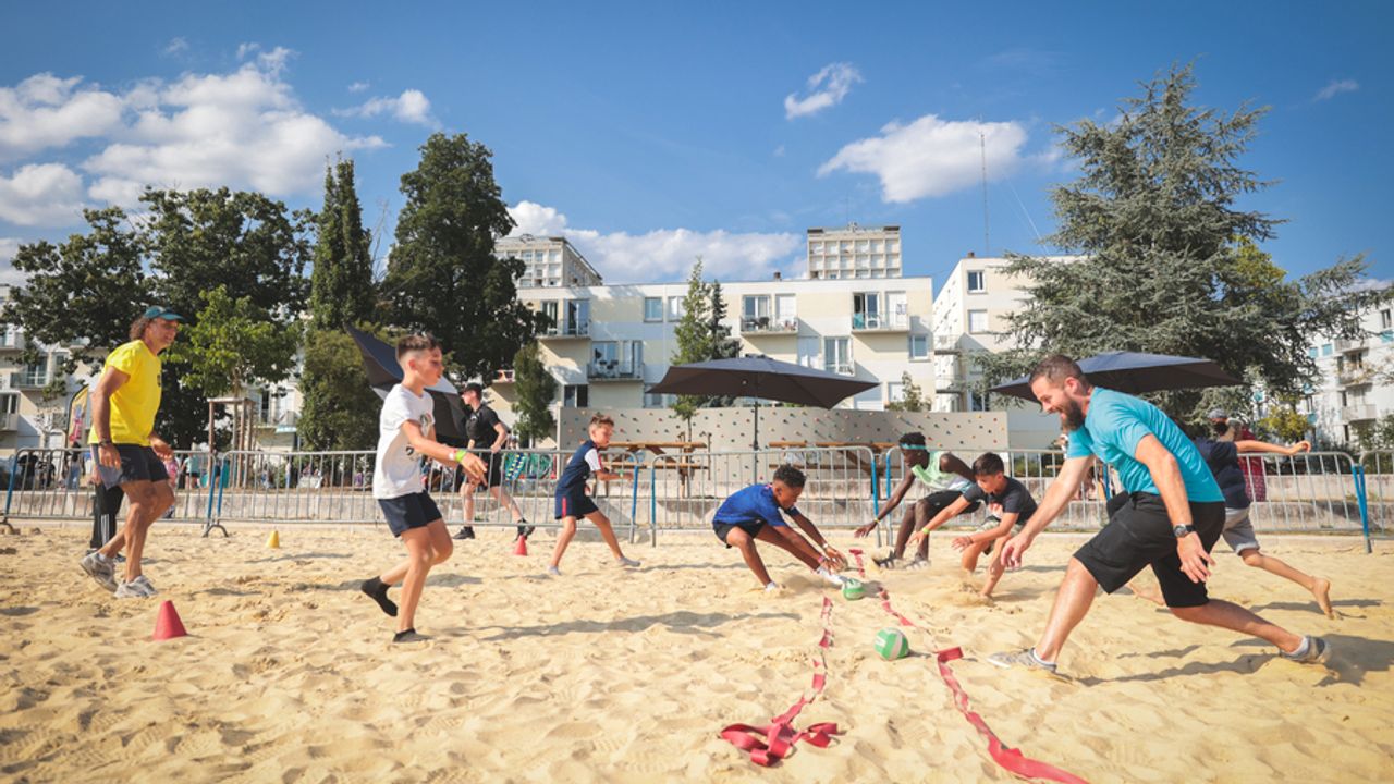 Un groupe de personnes (enfants, adolescents, adultes) joue à un jeu par équipe de 2 avec des balles sur la plage du parc du Berry dans le quartier de Villejean à Rennes. En arrière plan il y a des bancs et des parasols puis le mur d'escalade du parc du Berry et des arbres.