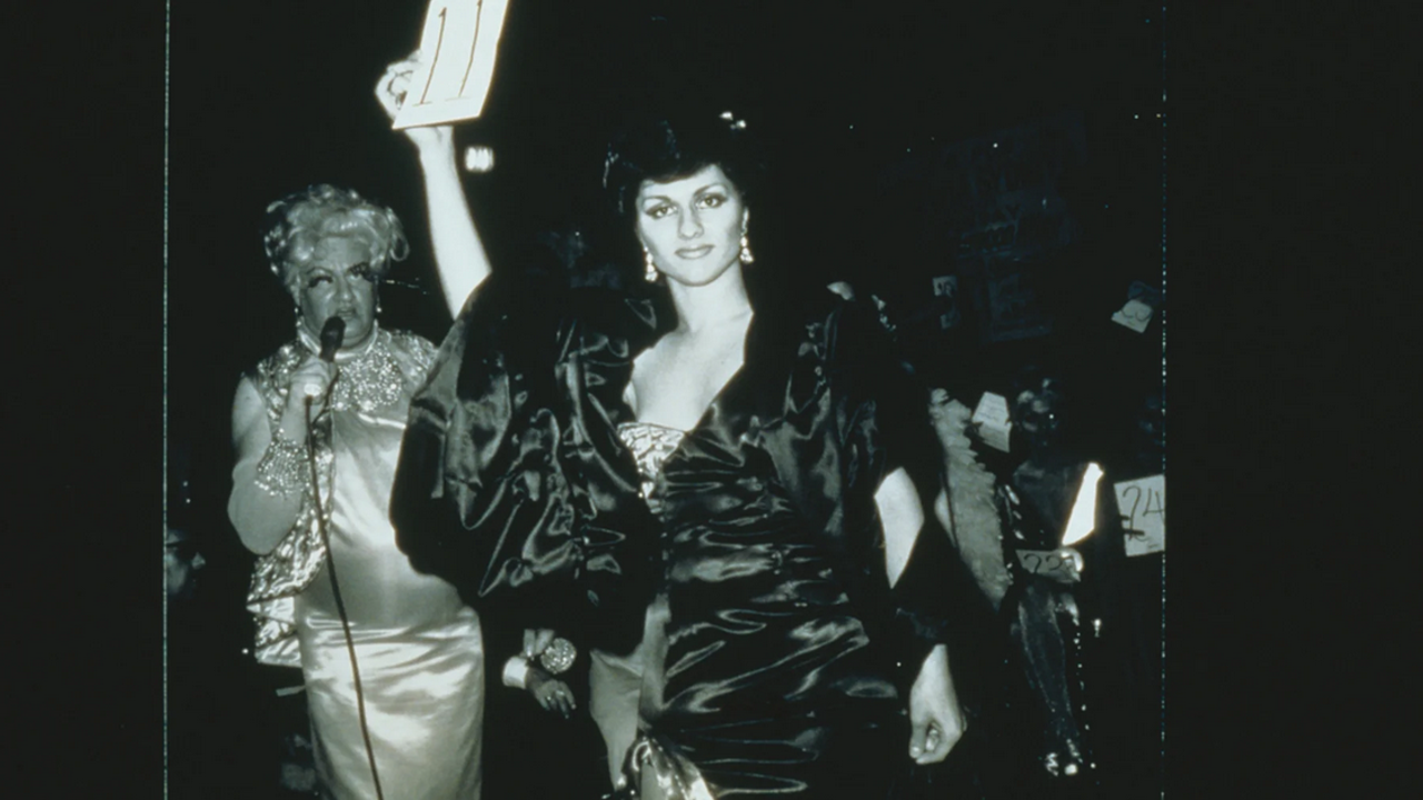 Sur la photographie de Nan Goldin, une femme parade et regarde fièrement l'objectif. Parée d'une robe volumineuse, elle brandit un papier où est inscrit le nombre 11.