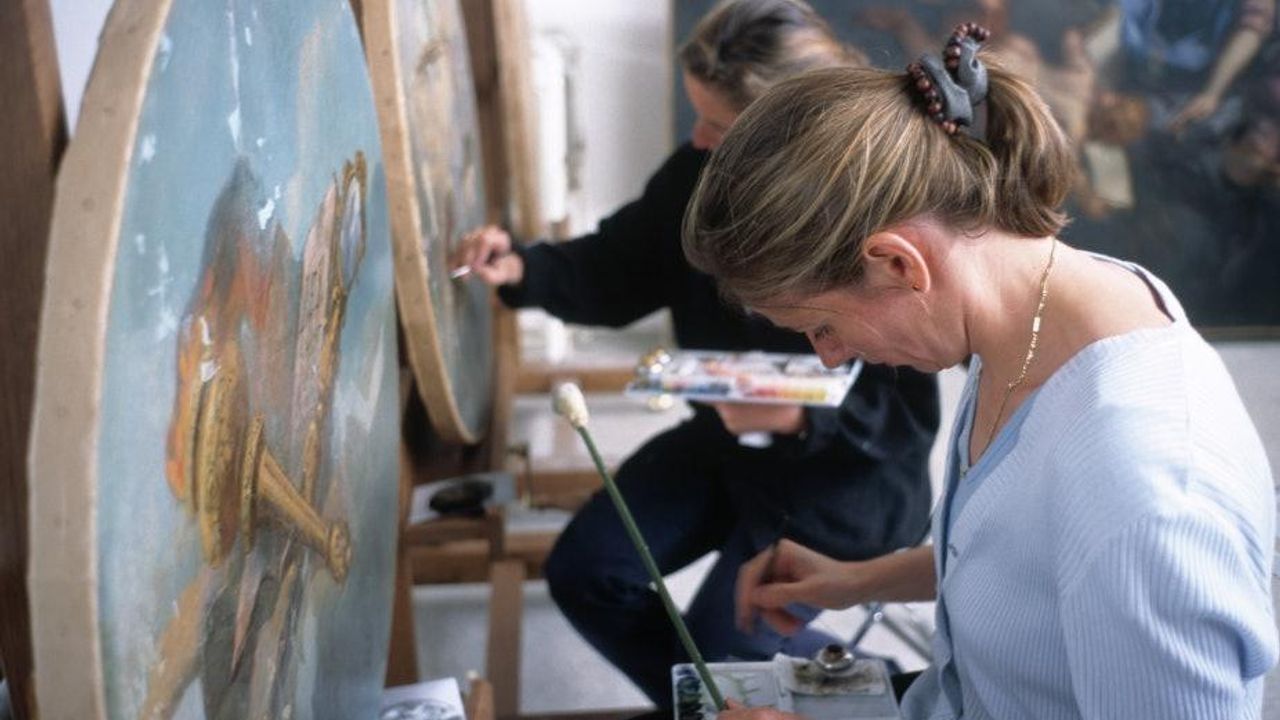 Deux personnes sont assises devant les toiles endommagées avec pinceaux et peinture afin de les restaurer.