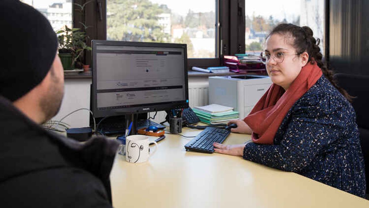 Une personne assise à un bureau devant un ordinateur face à une autre personne.