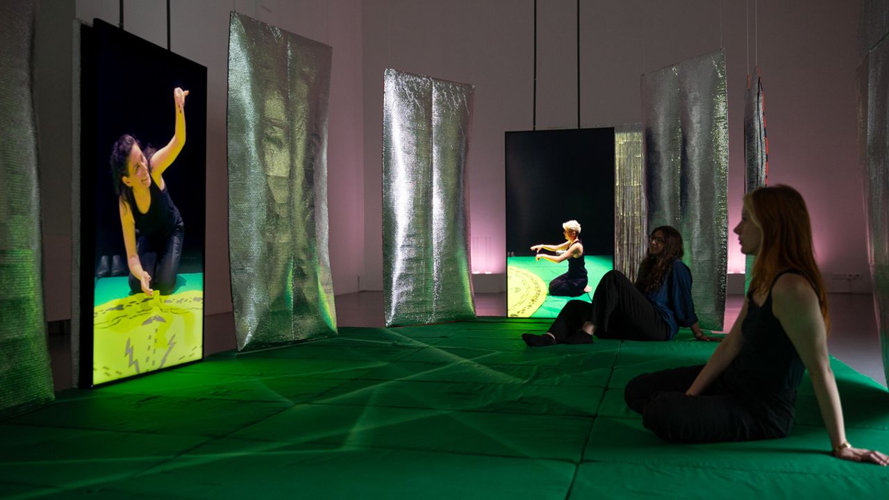 Deux personnes assises sur un tapis vert regardent deux vidéos diffusées sur de grands écrans verticaux.