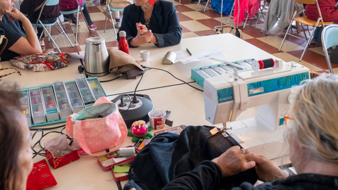 Au premier plan, des femmes reprisent des vêtements sur des machines à coudre. À l'arrière plan, les visiteurs patientent avec leurs objets usés pour une consultation avec les réparteurs.