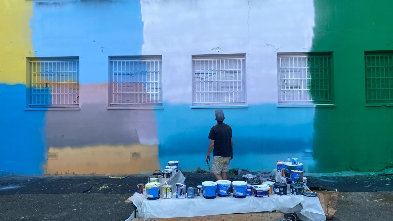 Une table avec des pots de peinture, un artiste qui regarde son oeuvre. Un mur de prison coloré.