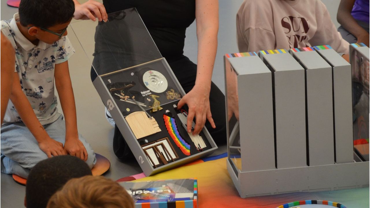 La médiatrice Amandine Braud présente aux enfants les oeuvres miniature du jeu de plateau collaboratif Rejouer La Criée aux enfants. Elles sont de différentes formes et couleurs, et sont inspirées de 40 oeuvres produites par La Criée depuis 40 ans.