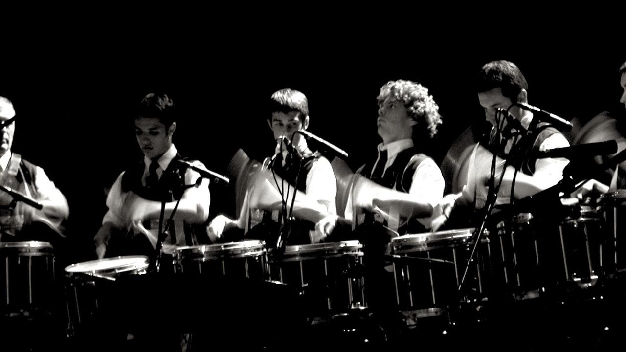 Sur scène, des musiciens alignés jouent des percussions.