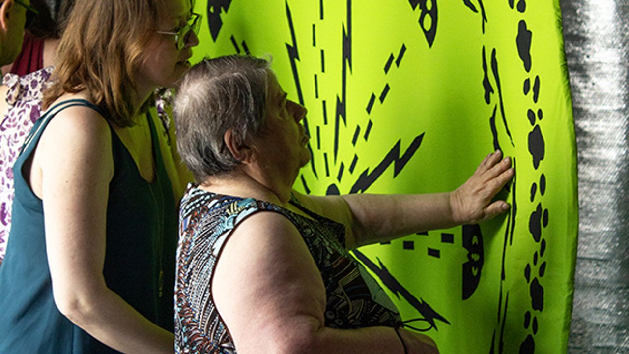 Une visiteuse accompagnée d'une médiatrice découvre l'oeuvre de Violaine Lochu à l'occasion d'une visite descriptive et tactile. L'oeuvre en question est un tapis vert anis en coton suspendu sur lequel sur cousu des motifs noirs semblant dessiner une chorégraphie ou une partition.