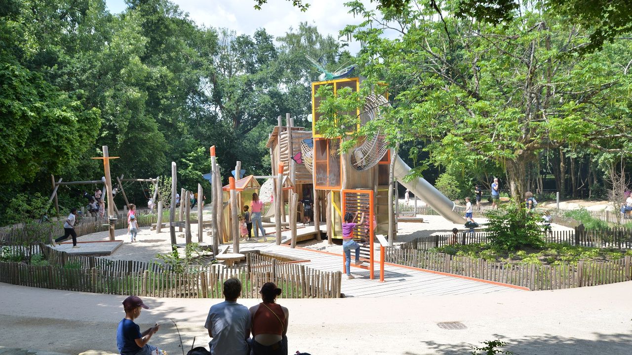En arrière plan, une aire de jeux pour enfants, on peut y grimper, faire du toboggan. Au premier plan un groupe de personne observe les enfants qui jouent sur l'aire de jeux.