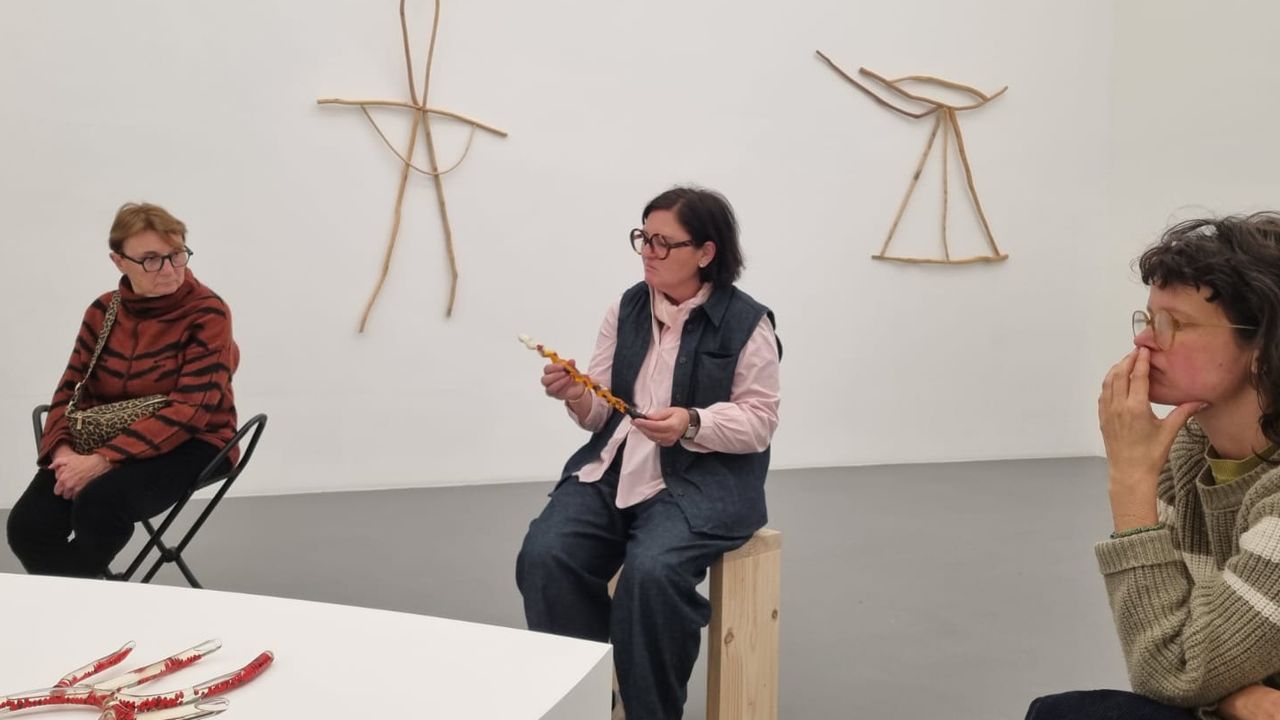 On voit trois personnes assises sur des sièges en bois. Dans le coin inférieur gauche on voit la pointe d'un grand socle en amande. Une sculpture composée de plusieurs tubes de verre déformés, assemblés et rempli d'une huile transparente ainsi que de graines rouges y est posée. Sur le mur derrière elles, on voit deux sculptures en bois assemblé. La femme au centre de la photographie tient dans ses mains et contemple un tube en verre dit "tremblé", lui aussi rempli d'huile et d'éléments naturels.