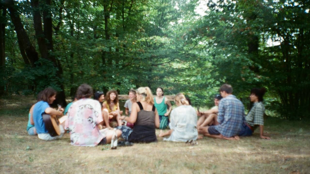 Group of people sitting on the grass in a circle by the forest singing and playing instruments