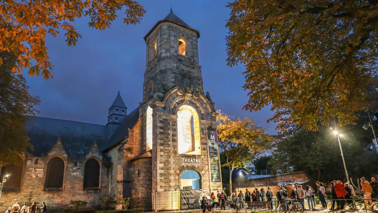 Vue de l’extérieur du Théâtre du Vieux Saint-Étienne situé dans une église. La nuit est tombée, l’église est éclairée et des personnes attendent d’entrer pour le festival Maintenant.