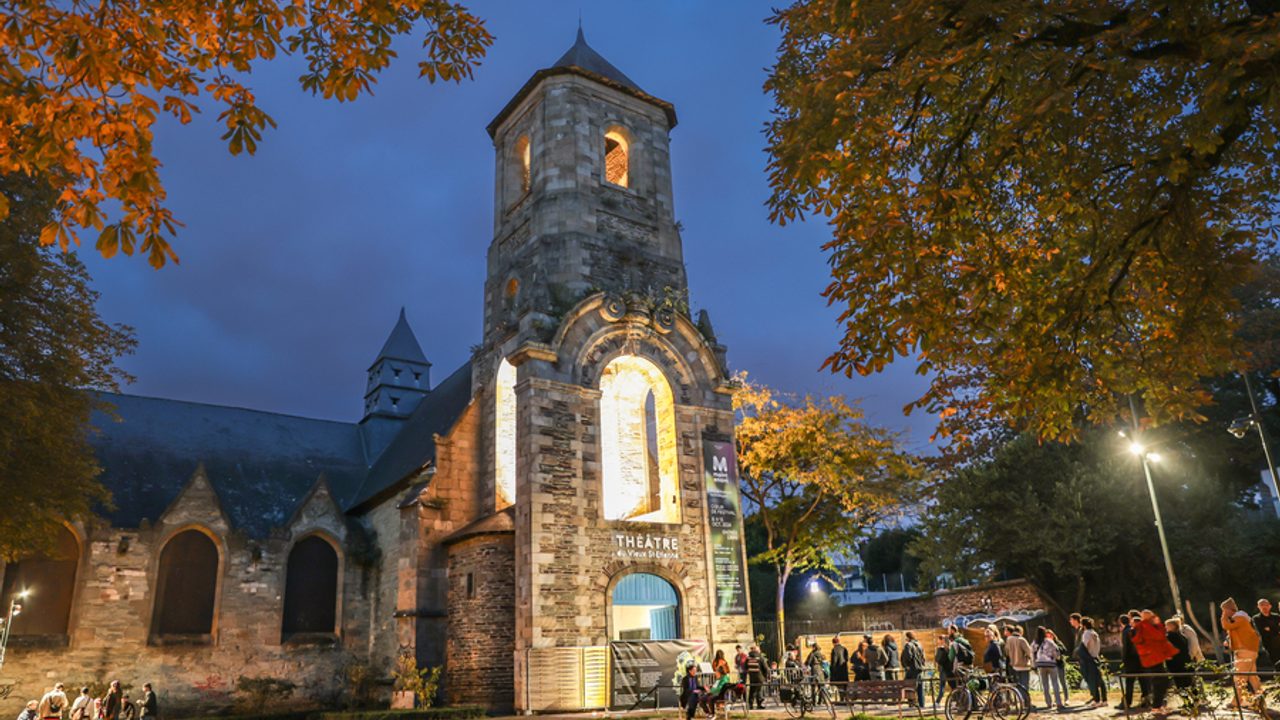 Vue de l’extérieur du Théâtre du Vieux Saint-Étienne situé dans une église. La nuit est tombée, l’église est éclairée et des personnes attendent d’entrer pour le festival Maintenant.