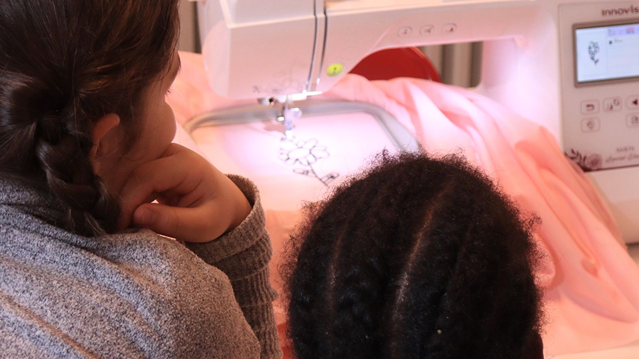 Deux élèves regardent la brodeuse reproduire un motif de fleur en fil noir sur un tissu rose.