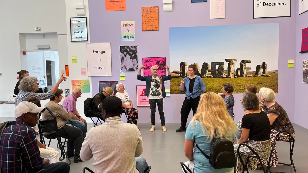 Au premier plan, des visiteurs assis sur des cannes-sièges, assistant à une visite à la Criée. Au second plan, une médiatrice explique l’exposition, accompagnée d’un traducteur en langues des signes.