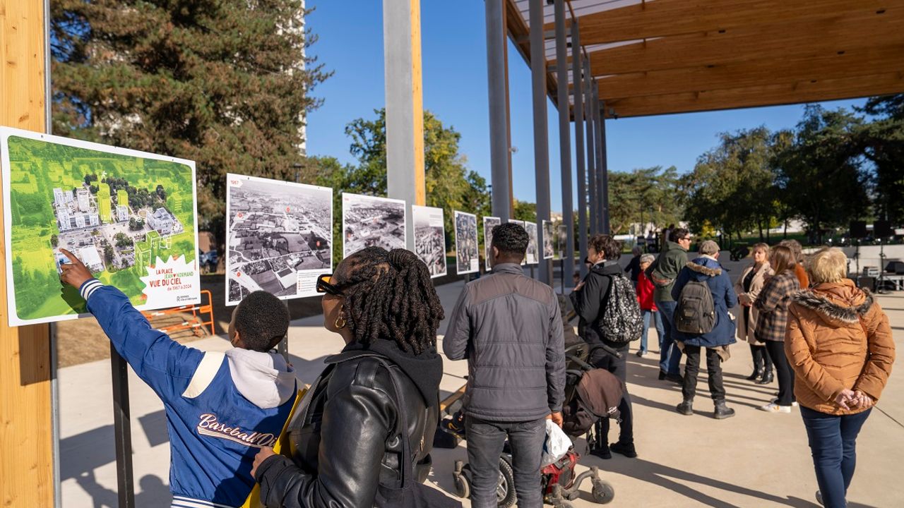 Vue d'une exposition de photographies sur la place Jean-Normand présentée sous la halle Agnès Sirop en octobre 2024. Des personnes regardent les photos, au premier plan, un enfant montre un point sur une photo colorée de la place Jean-Normand vue du ciel.