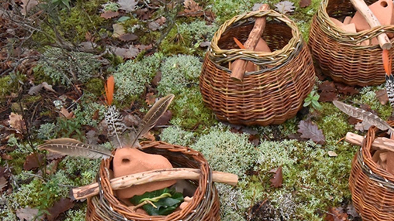 On voit plusieurs paniers en osier disposés sur un sol de forêt. A l'intérieur des paniers, on aperçoit des objets en céramique brute et ocre. Un objet dépasse du panier, il ressemble à une montagne et est orné de trois plumes.