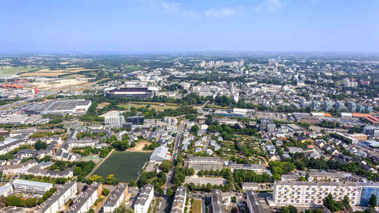 Vue aérienne de l'ouest de Rennes avec le quartier de Cleunay au premier plan, la Vilaine et le Roazhon Park en milieu de plan et en arrière-plan la campagne à gauche et le quartier de Villejean au centre et, en haut de l'image, le ciel.
