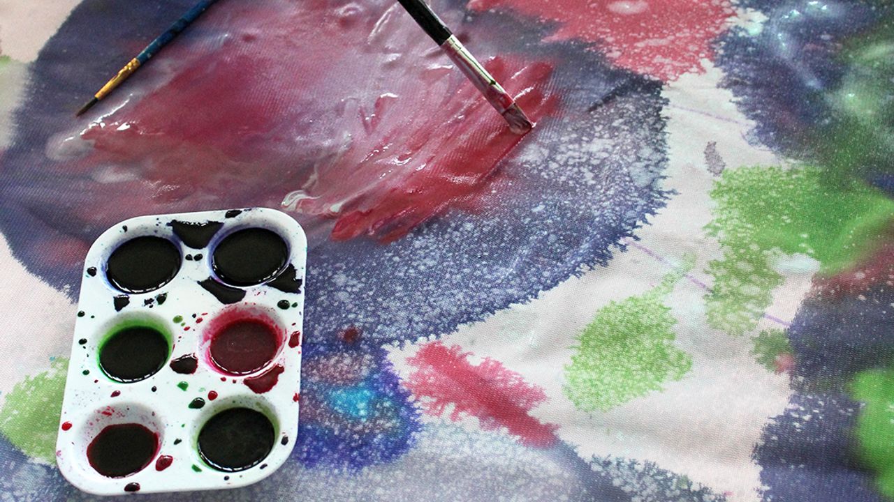 Un élève ajoute de la peinture rose sur son tissu déjà décoré de tâches et de motifs violets, verts et bleus.