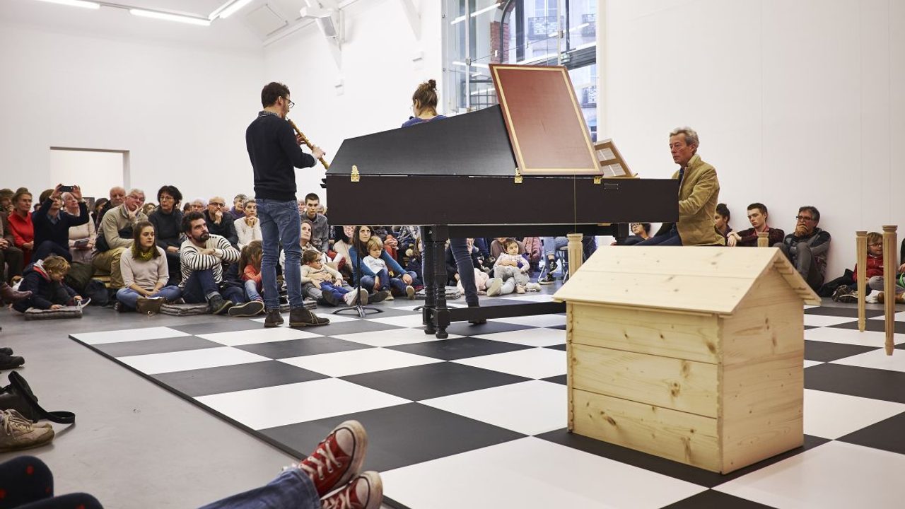 On voit un groupe de trois personnes jouer de la musique sur un grand damier pos au sol. Au premier plan, on voit une maison en bois sur une des cases du damier. Tout autour une foule assiste au concert.