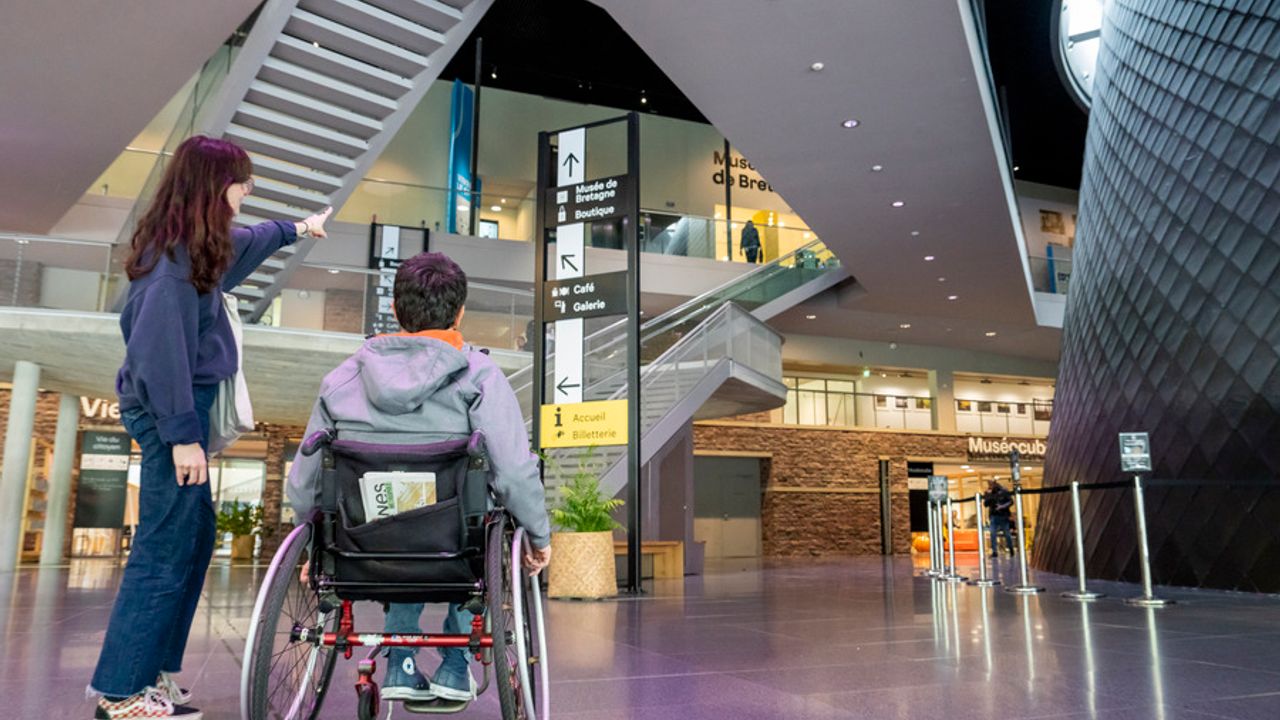 Au premier plan, une femme avec un homme en situation de handicap en fauteuil roulant. En arrière-plan, un panneau de signalétique clair dans le hall des Champs libres.